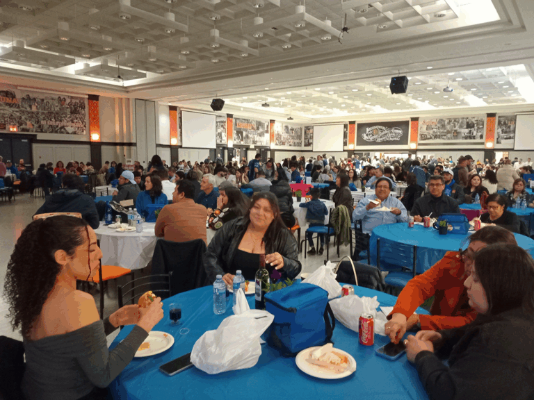 A large group of people gather in a spacious hall for the 2025 Historical Memory Festival . Tables covered with blue and white tablecloths fill the room as attendees eat, talk, and enjoy the evening. The walls display large black-and-white photos and posters celebrating community history.