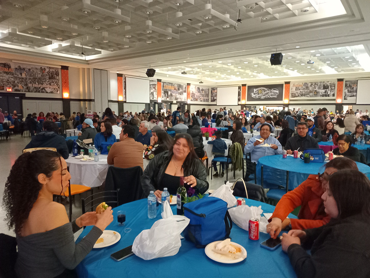 A large group of people gather in a spacious hall for the 2025 Historical Memory Festival . Tables covered with blue and white tablecloths fill the room as attendees eat, talk, and enjoy the evening. The walls display large black-and-white photos and posters celebrating community history.