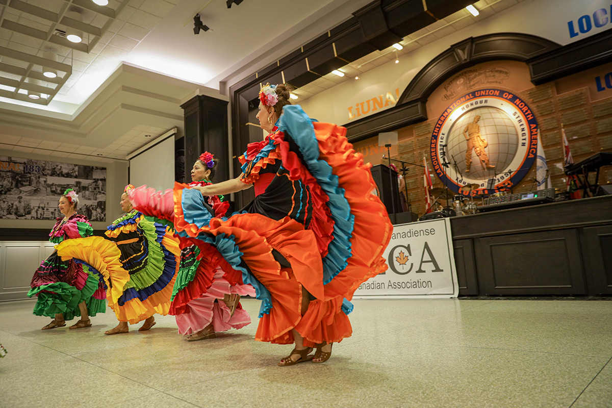 A group of women perform a traditional folkloric dance in brightly colored dresses during the 2023 Historical Memory Festival. They lift and swirl their skirts in unison on stage, celebrating Salvadoran culture and heritage at an indoor event hall.