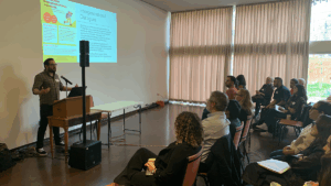 A speaker stands at a podium presenting to an audience during the “Academia Meets Community: Building Bridges Through Dialogue” conference. A slide titled “Intergenerational Dialogues” is projected on the wall as attendees listen attentively. The one-day event, co-organized by the Salvadoran Canadian Association and Western University, focuses on memory, justice, and education in postwar El Salvador.