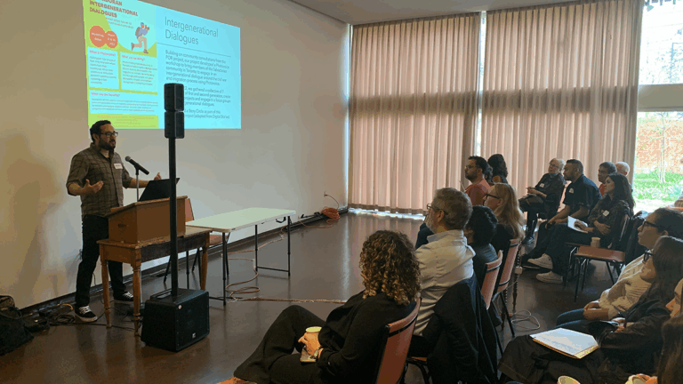 A speaker stands at a podium presenting to an audience during the “Academia Meets Community: Building Bridges Through Dialogue” conference. A slide titled “Intergenerational Dialogues” is projected on the wall as attendees listen attentively. The one-day event, co-organized by the Salvadoran Canadian Association and Western University, focuses on memory, justice, and education in postwar El Salvador.