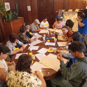 A group of people sit around a large wooden table participating in an embroidery workshop. They are stitching fabric pieces, chatting, and sharing materials like scissors, thread, and coffee cups in a warm, wood-paneled room.