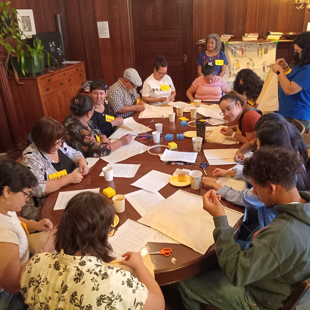 A group of people sit around a large wooden table participating in an embroidery workshop. They are stitching fabric pieces, chatting, and sharing materials like scissors, thread, and coffee cups in a warm, wood-paneled room.