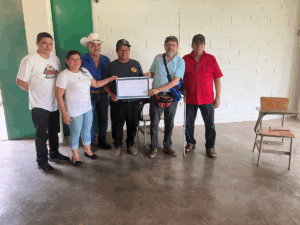 Six people stand indoors posing for a photo while holding a large check during a fundraising effort for Radio Farabundo Martí. The group includes community members and organizers supporting this radio station, which preserves historical memory and serves thousands across El Salvador.