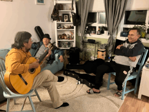 Three men sit together in a living room, two listening as one plays an acoustic guitar. Musical instruments and personal items fill the background shelves, creating a relaxed atmosphere. The photo was taken during interviews for the Virtual Museum of Migration Stories project.