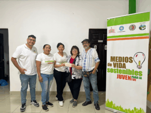A group of five people stand together smiling indoors beside a banner that reads “Medios de Vida Sostenibles para la Juventud.” Three people wear white shirts with the same logo, while one person hands a paper to another. The setting appears to be a community or workshop space with tiled floors and white walls.