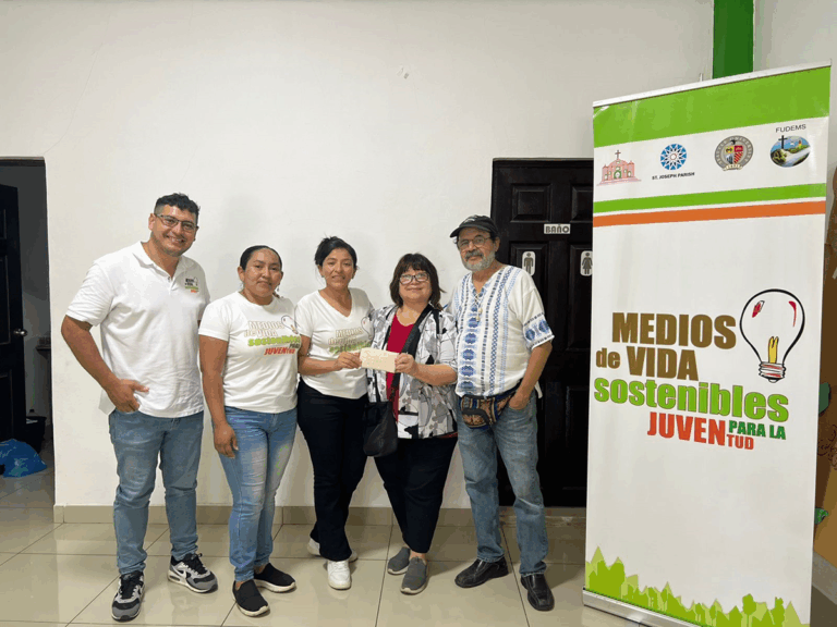 A group of five people stand together smiling indoors beside a banner that reads “Medios de Vida Sostenibles para la Juventud.” Three people wear white shirts with the same logo, while one person hands a paper to another. The setting appears to be a community or workshop space with tiled floors and white walls.