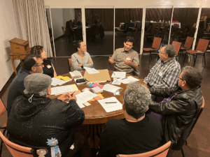 A group of eight people sit around a round table in discussion during a community meeting. Papers, notebooks, and pens are spread across the table as one person gestures while speaking. The session is part of ASALCA’s collaboration with Western University to launch The Book of the Salvadoran Diaspora, gathering insights from community leaders about migration and settlement experiences.