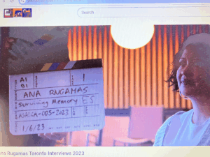 A film slate labeled “Ana Rugamas – Surviving Memory” is held up during an interview recording for the Virtual Museum of Migration Stories. A woman smiles softly under warm lighting, with vertical panels in the background. The project, organized by the Salvadoran Canadian Association (ASALCA) in collaboration with the University of Western Ontario, began its first series of interviews in Toronto.