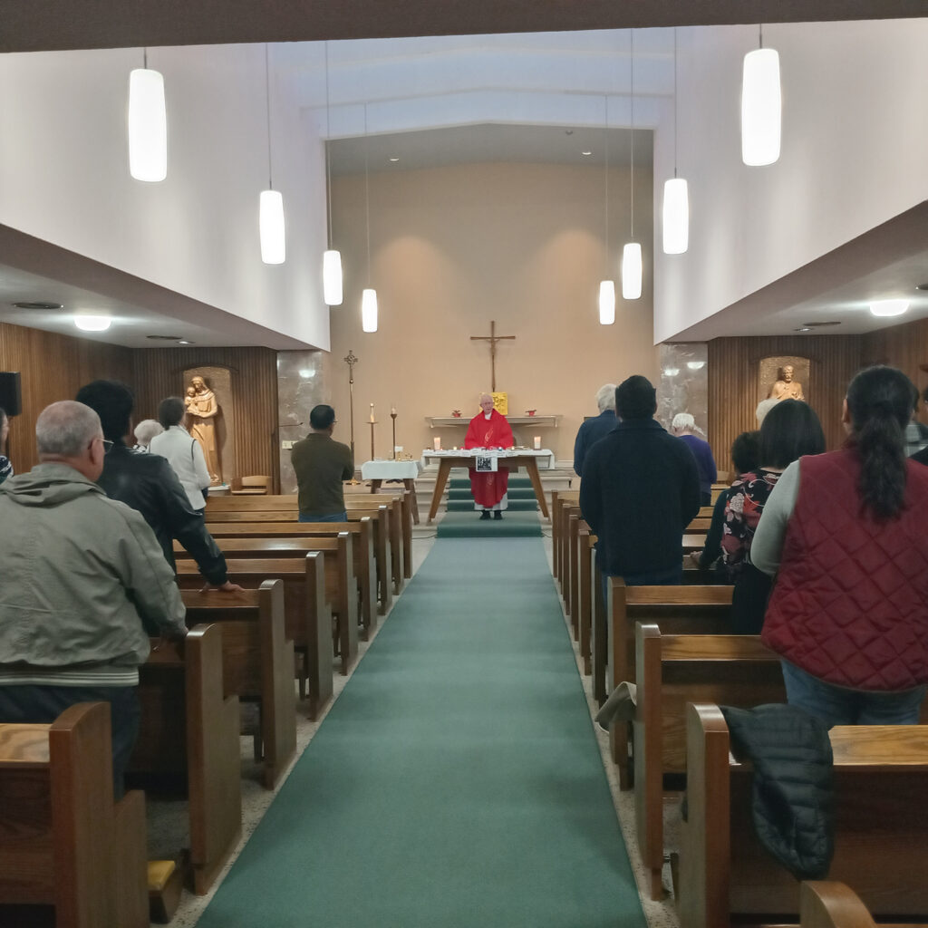 Congregation gathered in wooden pews during a church service. A priest in red vestments stands at the altar at the front of the church, with a cross on the wall behind him.