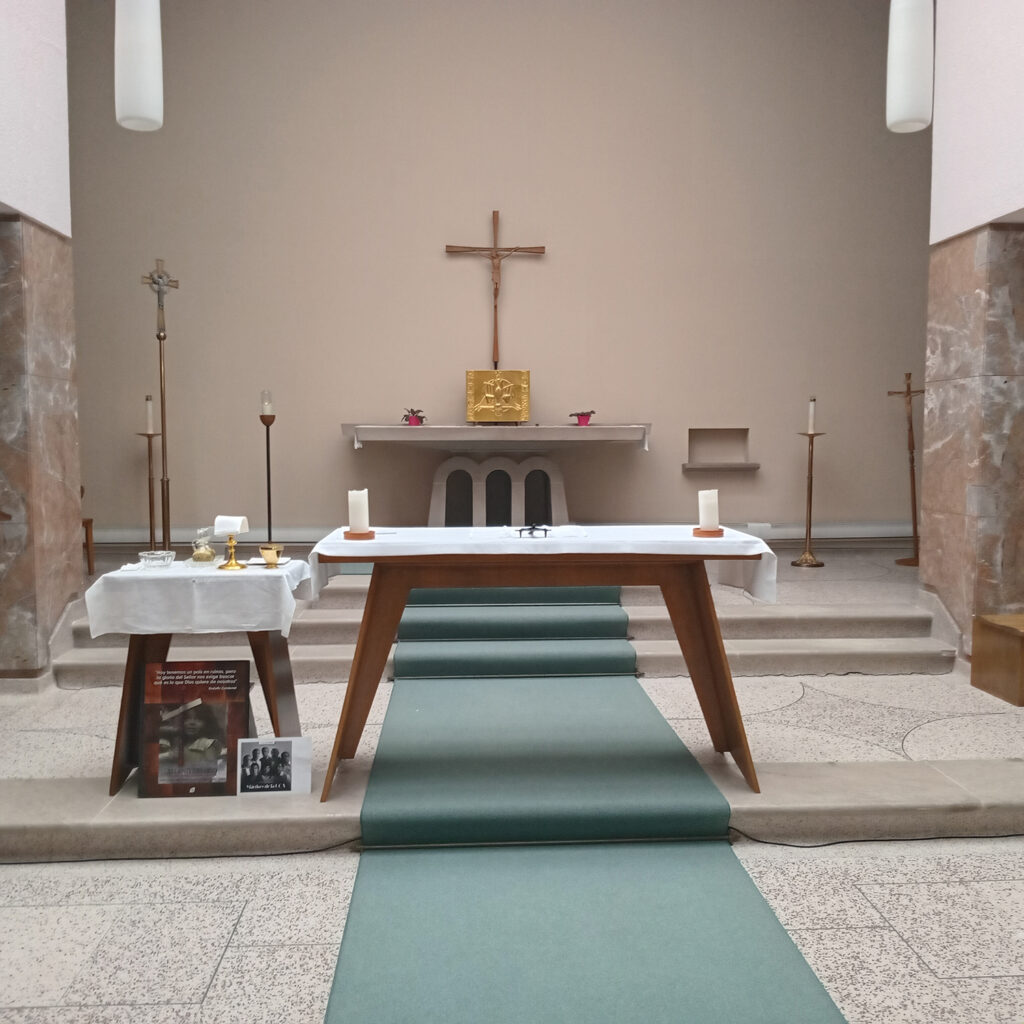 Close-up view of a church altar with white cloths, candles, a chalice, and framed photos arranged on small tables. A large cross hangs on the wall behind the altar.