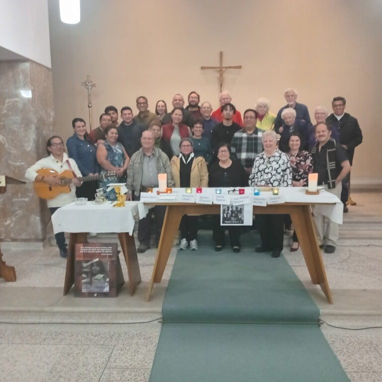 Large group photo inside a church. Around thirty people stand near the altar beneath a cross, smiling at the camera. A small table at the front holds candles, framed photos, and name cards honoring individuals.