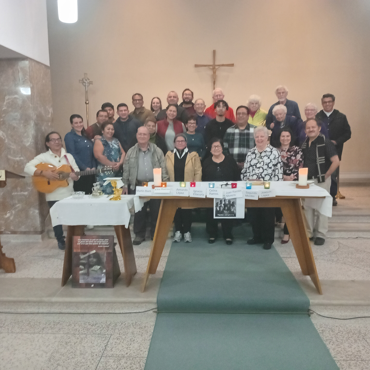Large group photo inside a church. Around thirty people stand near the altar beneath a cross, smiling at the camera. A small table at the front holds candles, framed photos, and name cards honoring individuals.