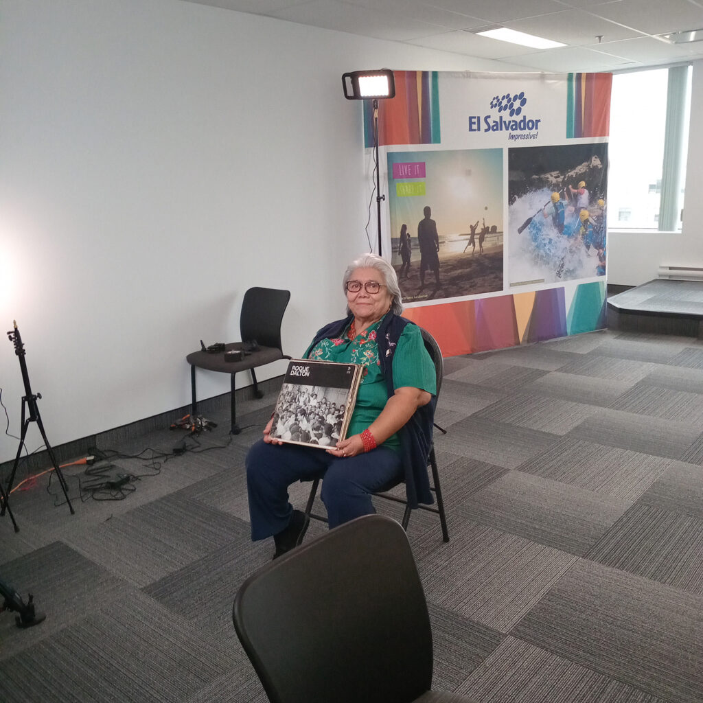 An older woman sits on a chair in a well-lit room holding a vinyl record with a black-and-white cover. Behind her is a colorful “El Salvador – Live it, Love it” backdrop and studio lighting equipment.