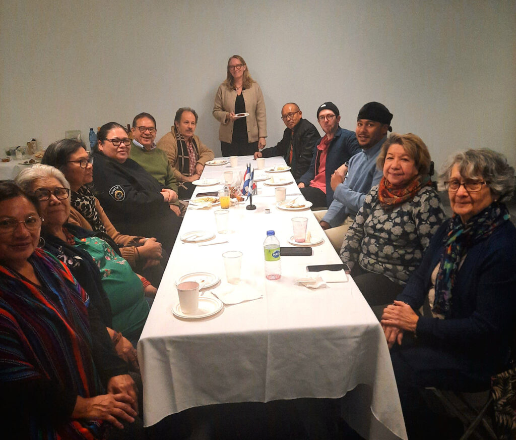 A group of twelve people sit around a long table covered with a white tablecloth during a gathering or meeting. Plates, cups, and small flags are on the table, and everyone is smiling toward the camera.