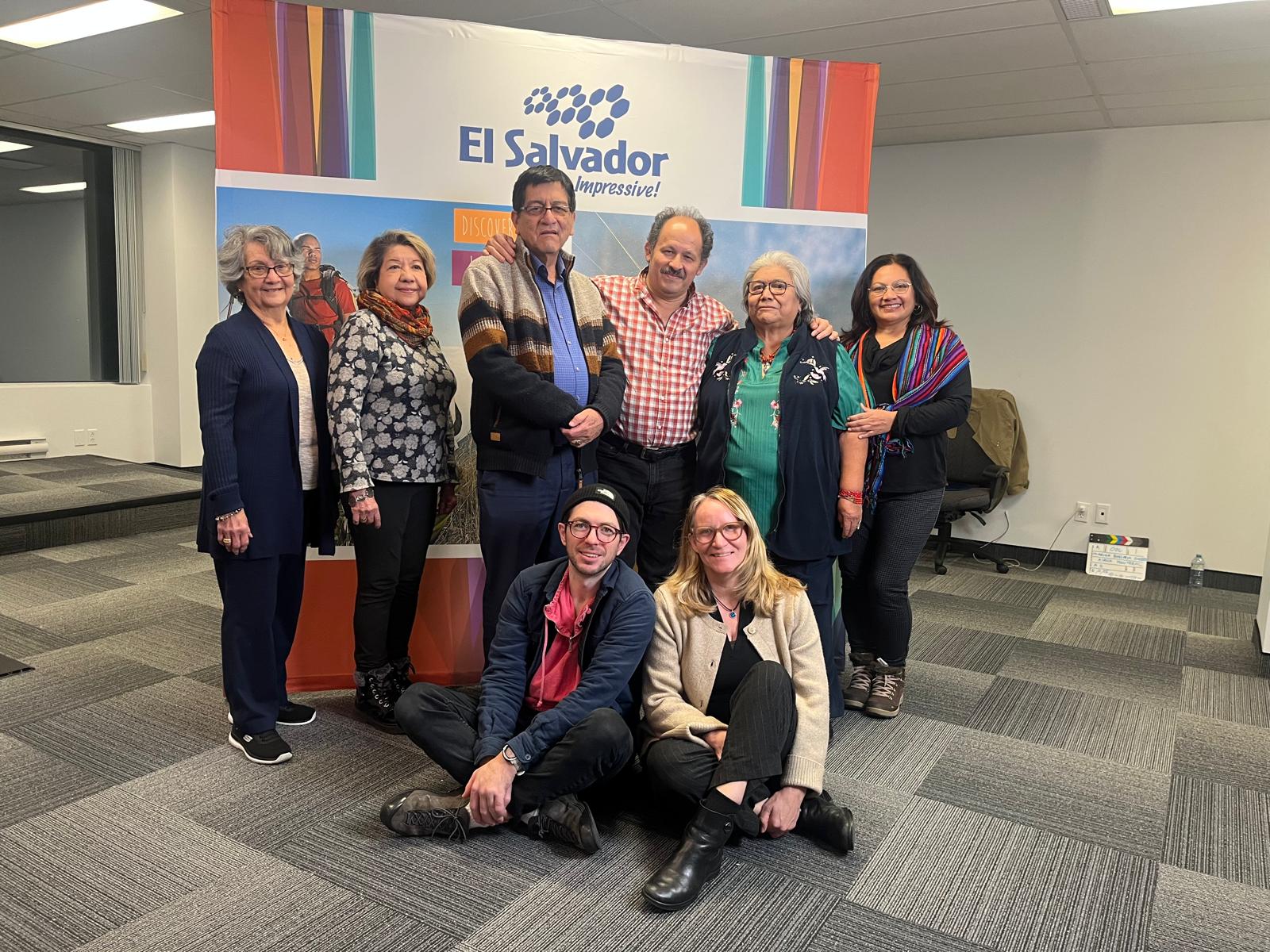 Eight adults stand together in front of a colorful “El Salvador – Impressive!” backdrop, smiling at the camera. Two additional people sit cross-legged on the floor in front of the group.