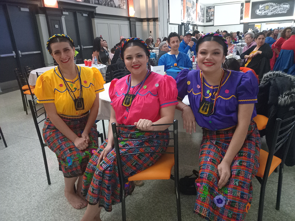 Three women dressed in vibrant traditional Central American attire—embroidered blouses in yellow, pink, and purple with plaid skirts—smiling while seated at a community cultural event, with attendees gathered at tables behind them.