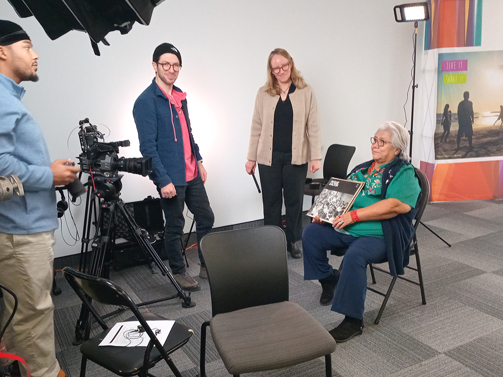 A woman in traditional embroidered clothing sits in a chair holding a photo album while being filmed by a crew in a studio setup, with cameras, lights, and two crew members standing nearby.